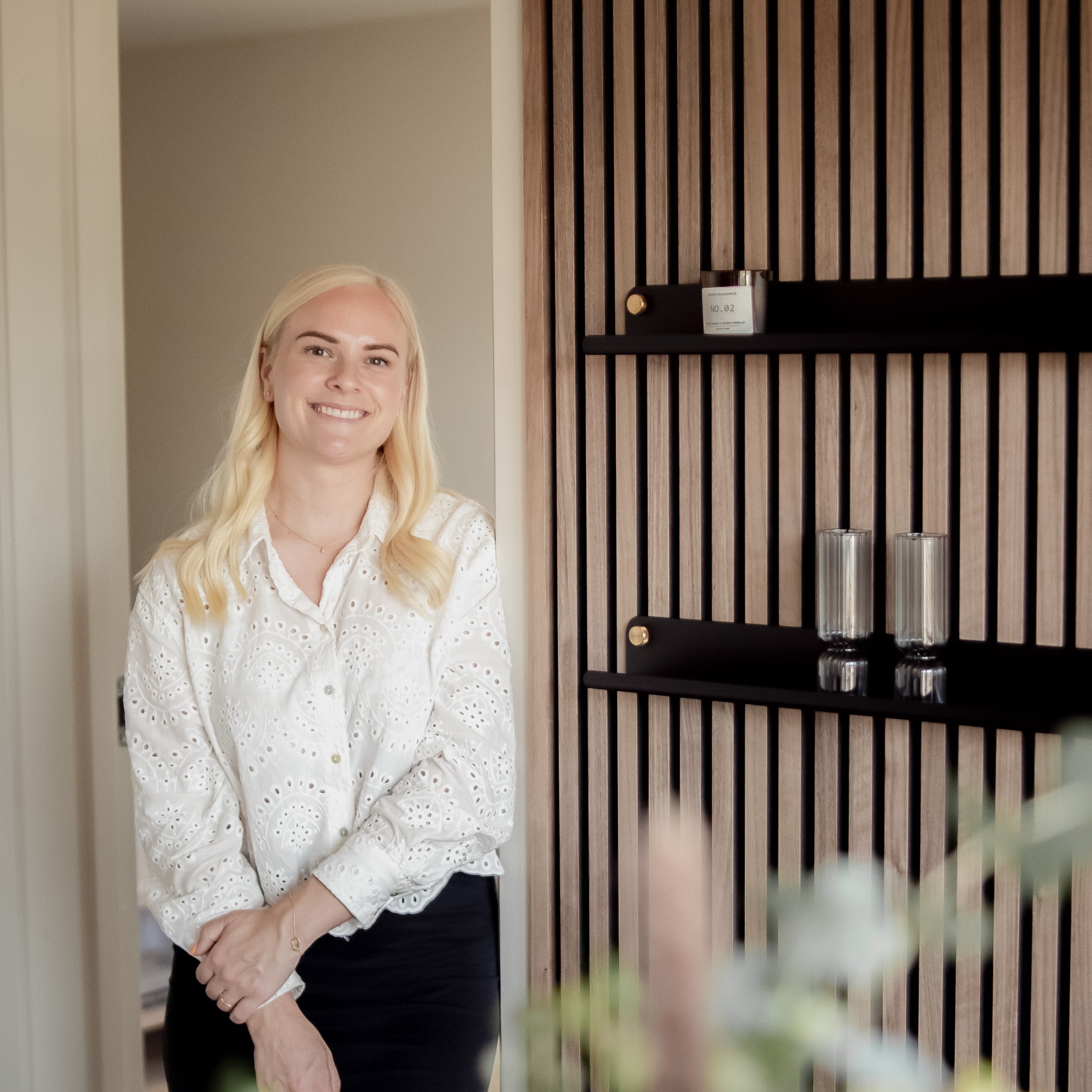 A woman with long blonde hair in a white blouse stands indoors beside a modern Akupanel acoustic wood panel wall with minimalist shelves.
